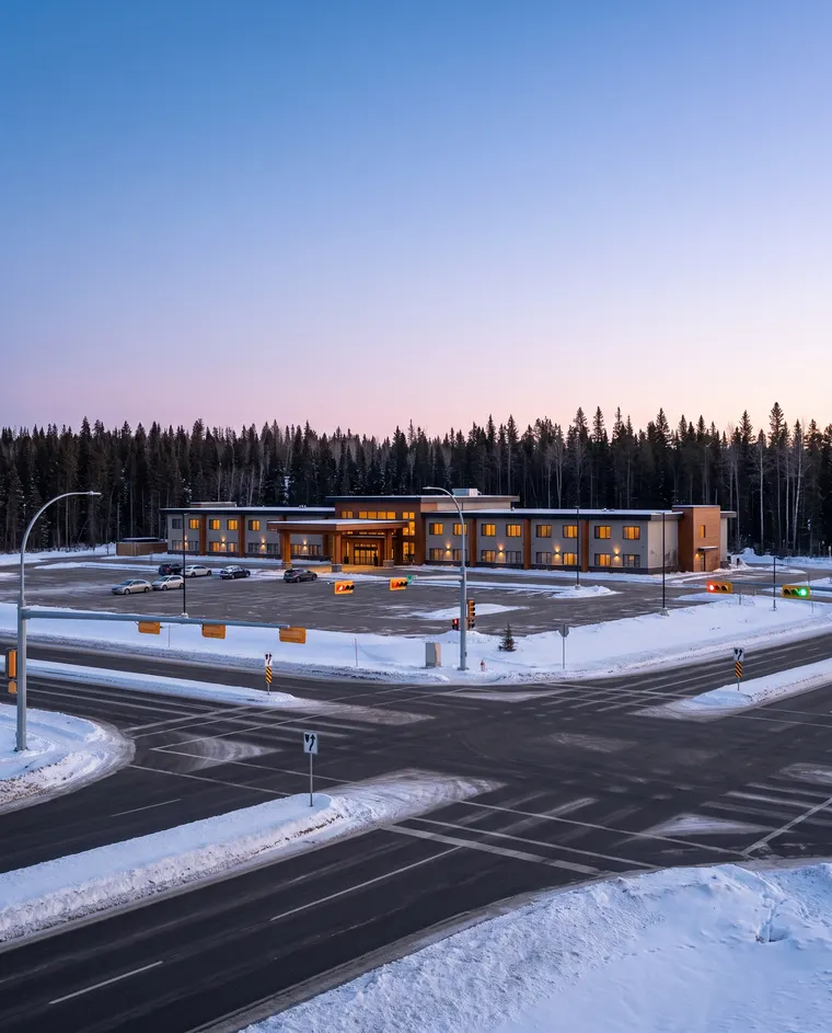 Generated illustration: a snow-ploughed highway intersection at winter dusk, a long low hotel and casino building with lit windows beyond a parking lot, and dark spruce forest behind; not a photograph of the casino.